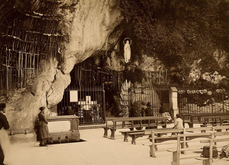Sepia photograph of a rock grotto hung with crutches, with a statue of Mary in a niche, an iron railing, and rows of benches