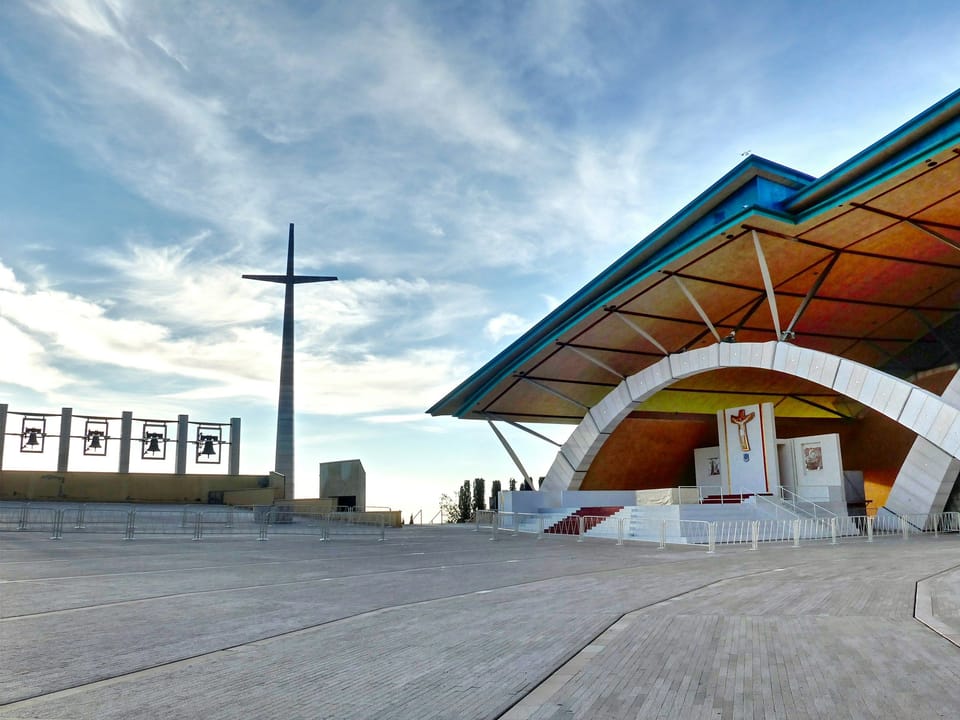 An outdoor altar under a broad sloping roof, with a tall cross and a row of bells beside a stone plaza