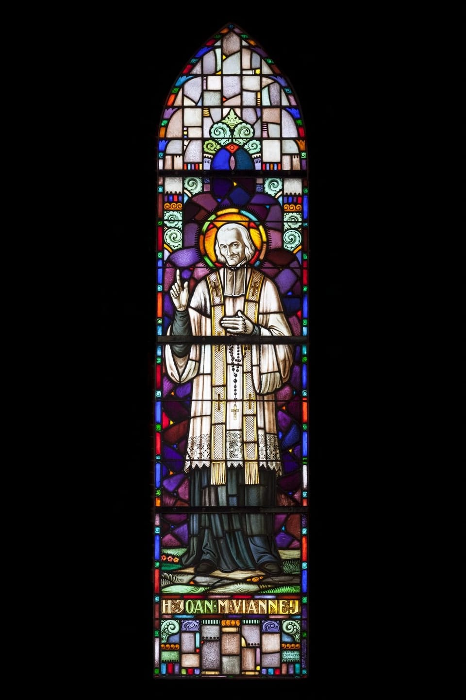 Stained-glass window of a priest in a white surplice raising one hand, with the name H. Joan. M. Vianney beneath